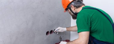 Bir elektrikçi daireye prizler takıyor. Turuncu kasklı ve tulumlu bir adam evde elektrik üretiyor..