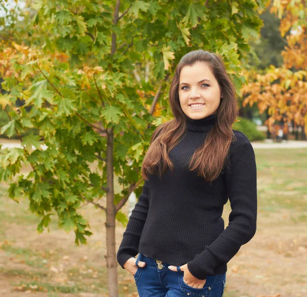 Atractiva chica sonriente en suéter negro de pie debajo de un árbol i — Foto de Stock