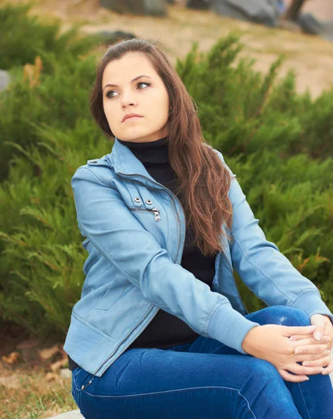 Attractive young girl in a blue jacket, sitting on a park bench — Stock Photo, Image