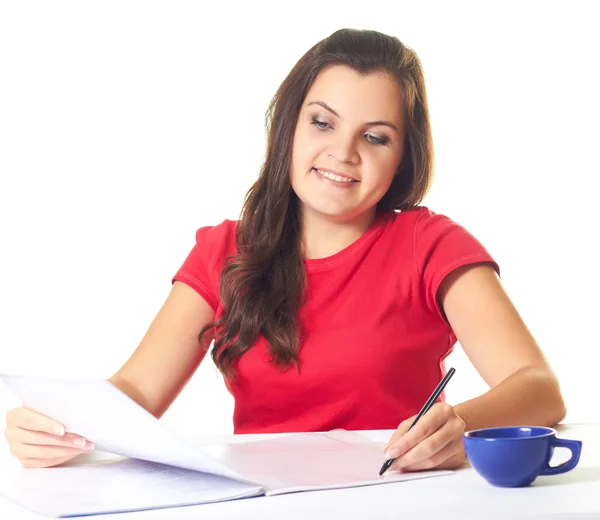 Привлекательная девушка в красной рубашке сидит за столом и пишет: Стоковое Изображение