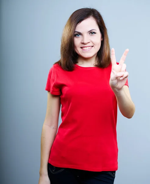 Usměvavá mladá žena v červené košili. ukazuje symbol vítězství. — Stock fotografie