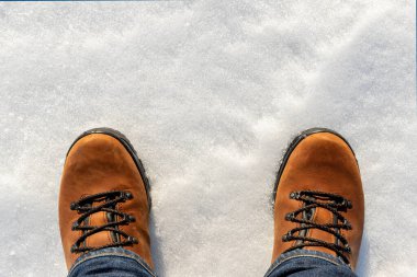 Üst tarafta kot pantolon giymiş erkek bacakları ve beyaz buzlu kar yüzeyinde izole edilmiş kahverengi deri çizmeler var. Su geçirmez ayakkabılar. Kış yürüyüşü ve doğa yürüyüşü kavramı