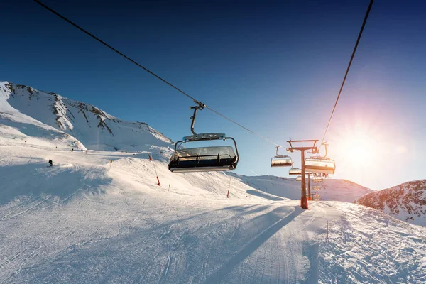 Teleférico vacío en la montaña montañosa alpina estación de invierno en la tarde soleada brillante. Telesilla de esquí vía cable con la gente disfruta del esquí y el snowboard. Cielo atardecer retroiluminado brillando en el fondo — Foto de Stock