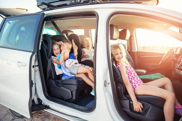 Group of four cute adorable little happy caucasian children enjoy having fun sit in minivan going to sea beach road trip on hot summer day. Kids in swimsuit in car with side open door against sun — Stock Photo, Image