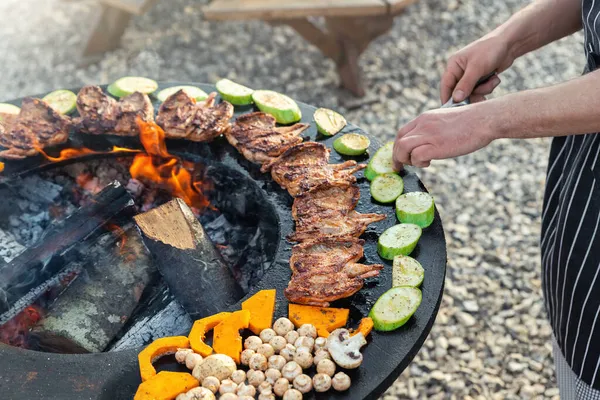 Close-up detail view of chef flipping by tongs tasty crispy marinated quails and vegetables grilled at round steel iron firepit hearth table surface brazier with burning firewoods. Barbecue yard home — Stock Photo, Image