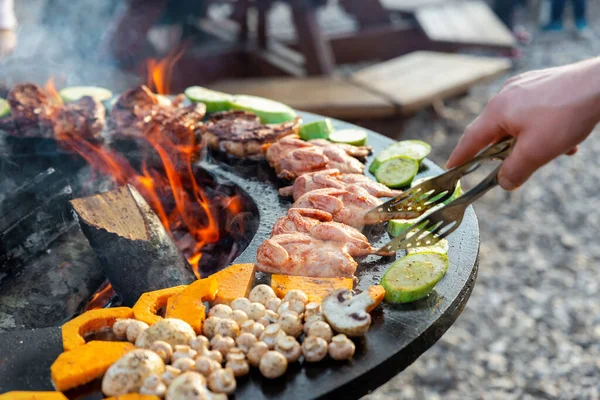 Close-up detail view of chef flipping by tongs tasty crispy marinated quails and vegetables grilled at round steel iron firepit hearth table surface brazier with burning firewoods. Barbecue yard home — Stock Photo, Image