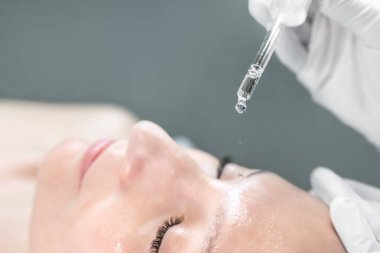 Yakın plan güzellik uzmanı koruma eldivenleri verdi. Yaşlanma karşıtı prosedürler uyguluyor. Güzellik kliniğindeki genç çekici bayan müşteriyle organik kozmetik losyon sürüyor. Cilt bakımı kavramı