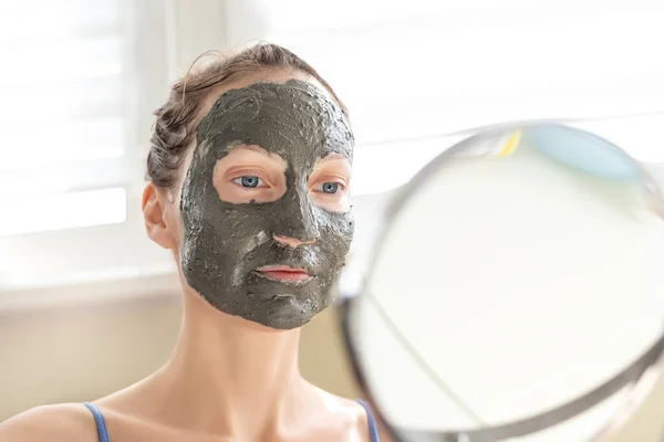 Genç, mutlu, beyaz bir kadın gündüz vakti evde cam ve aynaya karşı doğal gri kil yüz maskesi takıyor. Kadın yüz temizleyici, nemlendirici tedavi uyguluyor. — Stok fotoğraf