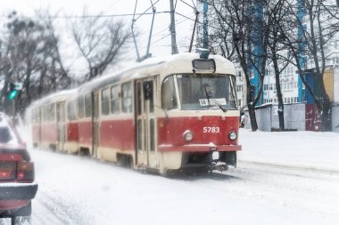 Soğuk kış gününde yoğun kar fırtınasında trafik varken eski model tramvayın karlı şehir sokaklarını sürüşünün manzarası. Işınlama, soğuk mevsim havası. Kiev, Ukrayna - 02 Şubat 2021
