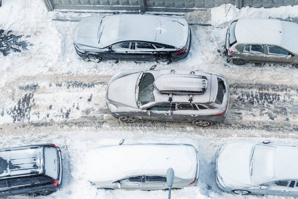 Top above aerial view of modern suv crossover car with roof rack box trunk driving through vehicles parked on city street covered by deep snow on clod frosty winter day. Apartment building driveway