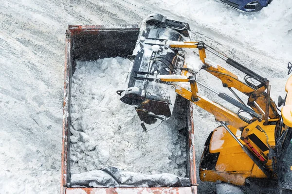 Máquina cargadora de tractores cargando nieve sucia en un camión volquete. Limpieza de la calle de la ciudad, la eliminación de la nieve y el hielo después de fuertes nevadas y ventiscas. Arado de nieve al aire libre acera pavimento limpio camino de entrada — Foto de Stock