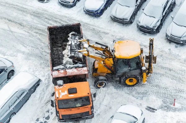 Máquina cargadora de tractores cargando nieve sucia en un camión volquete. Limpieza de la calle de la ciudad, la eliminación de la nieve y el hielo después de fuertes nevadas y ventiscas. Arado de nieve al aire libre acera pavimento limpio camino de entrada — Foto de Stock