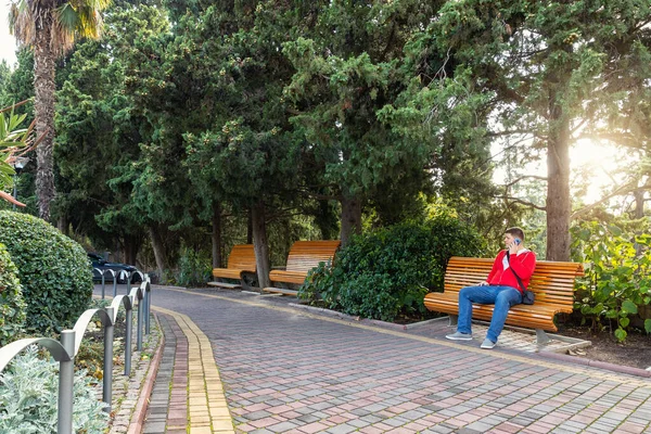 Jonge volwassen mannelijke persoon in casual dragen zitten op de bank in de stad straat groene park tuin met behulp van smartphone. Man rustend in Parkland pratend per telefoon — Stockfoto
