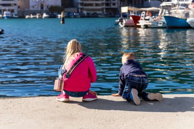Arkaya bakan iki sevimli küçük meraklı kardeş deniz kıyısının yakınındaki beton set iskelesinde oturup yat teknesi marinasını izliyorlar. Çocuklar yaz günü dışarıda oynamaktan zevk alırlar.