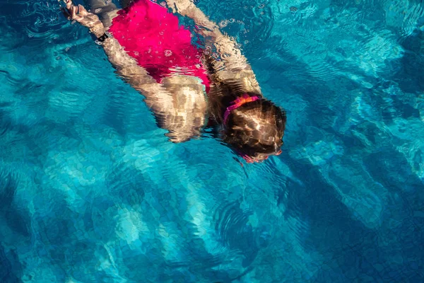 Vista superior de la niña linda en traje de baño de color rosa deportivo y gafas de buceo bajo el agua en agua azul claro de la piscina cubierta a la hora de la noche oscura. Niños deportes acuáticos actividades saludables — Foto de Stock