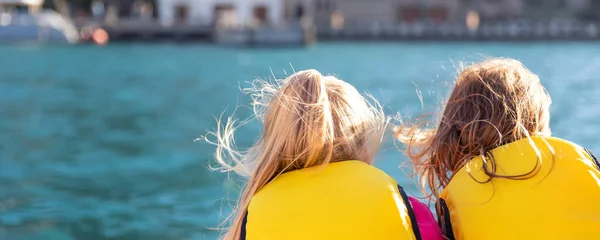 Vista trasera dos lindos rubios felices emocionados sonrientes amigas caucásicas usan chaleco salvavidas disfrutan navegando en el borde del barco a motor mar contra el sol brillante de agua azul. Vacaciones de verano recreación — Foto de Stock