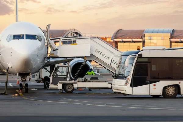 Front view big modern passenger aircraft approach boarding witth stairs vehicle and shuttle bus against airport building morning evening warm sun light time. Plane runway service maintenance handling