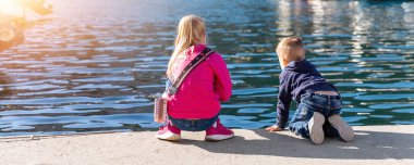 Arkaya bakan iki sevimli küçük meraklı kardeş deniz kıyısının yakınındaki beton set iskelesinde oturup yat teknesi marinasını izliyorlar. Çocuklar yaz günü dışarıda oynamaktan zevk alırlar.