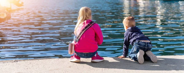Voltar ver duas crianças engraçadas pouco curiosos irmãos sentados no cais de diques de concreto perto do banco do lago do mar e assistindo navio de peixe iate marina. As crianças gostam de se divertir jogando ao ar livre no dia de verão — Fotografia de Stock