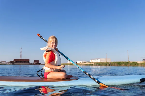 Cute pequena menina loira caucasiana desfrutar de se divertir sentado no surf prancha de água doce lago ou rio quente dia ensolarado. Verão ao ar livre criança esporte saudável atividades recreativas — Fotografia de Stock