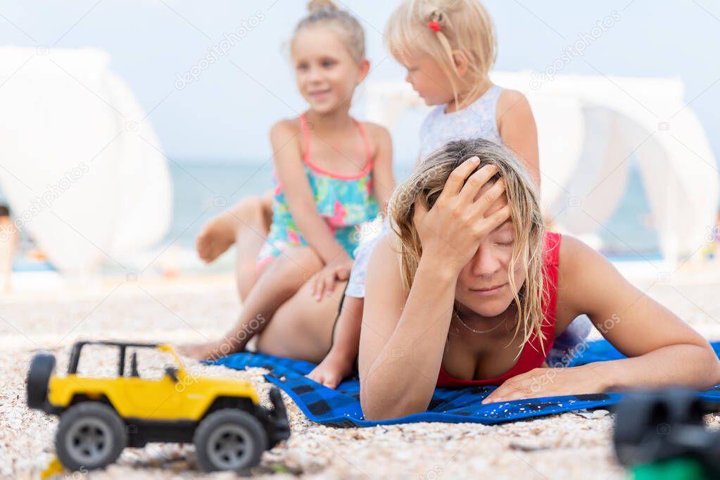 Two cute little sibling girls enjoy having fun playing sitting on tired exhausted mothers back at sea ocean beach. Frustrated mom make face palm gesture. Vacation family small kids trouble concept
