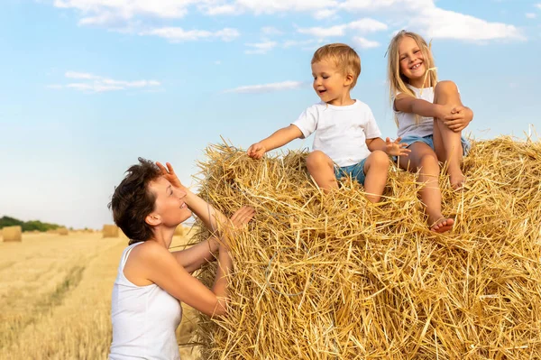 Jovem adulto atraente mãe bonita com filho pequeno e filha gostam de se divertir brincando sentado perto de fardo de feno dourado no campo colhido de trigo perto da fazenda. Crianças felizes na paisagem rural — Fotografia de Stock