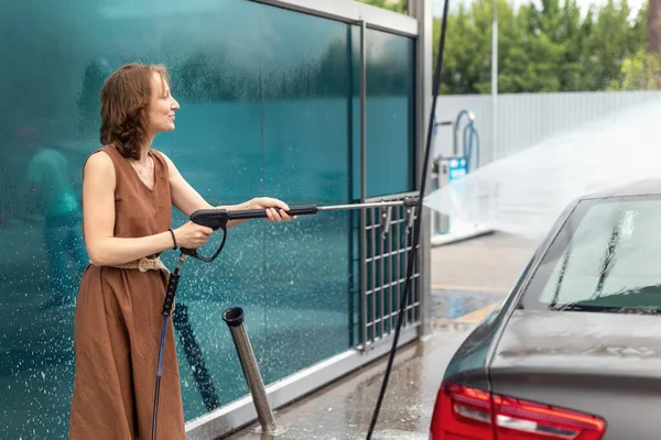 Vista lateral jovem mulher adulta lavar carro com bomba de equipamentos de água de alta pressão auto-serviço ao ar livre no dia de verão ventoso brilhante. Veículo coberto espuma shampoo químicos detergentes carwash auto-serviço — Fotografia de Stock