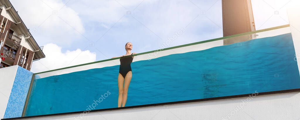 Young adult caucasian sporty slim smiling relaxing woman enjoy stand in pool with clear blue water and trasparent wall edge against sky at luxury spa resort. Healthy lifestyle and wellness concept
