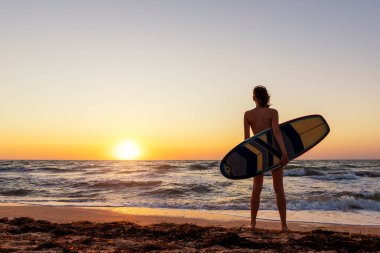 Okyanus kıyısında, güneşin doğuşuna ya da günbatımına karşı sörf tahtası silueti olan genç, sıska, sportif sörfçü kız. Spor sağlıklı kaygısız özgürlük yaşam tarzı tatil konsepti