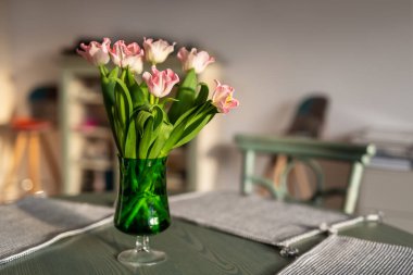 Masanın üzerinde yeşil cam vazodaki güzel pembe lale buketi kumaş masa örtüsü peçetesi ve sıcak gün batımında ev içi sıcak güneş ışığı. Çiçek açan oturma odası dekorasyon tarzı.