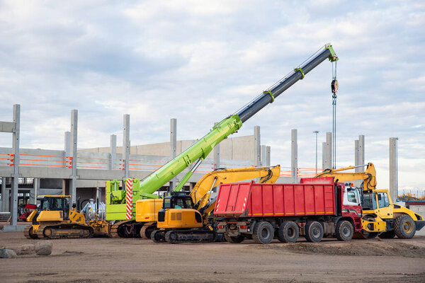 Many different multiclored colorful heavy industrial machinery equipment at construction site parking area against warehouse building city infrastructure development. Commercial vehicles rental sale.