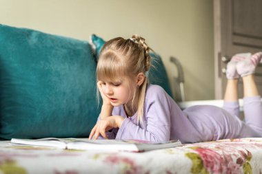 Gülümseyen küçük sevimli sevimli sevimli, mutlu, beyaz saçlı kız kanepeye uzanmış masal kitabı okumaktan zevk alıyor. Küçük kadın çocuk okuma, rüya görme ve hayal kurma gelişimi.