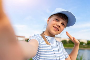 Küçük şirin sarışın beyaz kızın portresi mavi başlık takıyor ve yaz günü sahilde dinlenirken selfie fotoğraf makinesi çekerken eğleniyor. Çocuk blogları ve video yayınları.