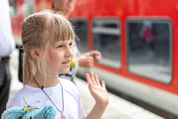 Portait of cute beautiful caucasian little blond kid girl looking waving hand hello goodbye gesture farewell at railway platform station agains red wxpress train. Child meeting friend at railroad.