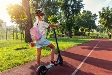 Profil portresi, güneşli bir günde açık havada elektrikli scooter şehir parkına binerken kask takan sevimli sarışın küçük beyaz kız portresi. Dışarıda sağlıklı çocuk aktiviteleri.