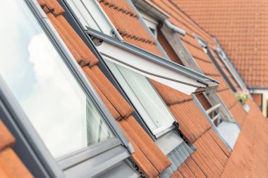 Open ventilation waterproof rooftop window exterior against sunny sky light. Velux style roof with red brick tiles. European city street attic mansard modern roofwindow service, install maintenance. clipart