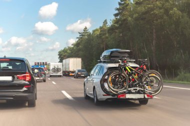 Arka plan modern beyaz aile arabası monte edilmiş çatı kutusu bagajı ve bisiklet kuyruğu taşıyıcısı Avrupa karayolunu mavi gökyüzüne doğru sürüyor. Hayat tarzı seyahat macera yolculuğu konsepti.