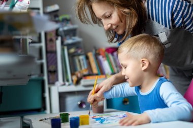 Young adult mom and cute little blond kid boy enjoy have fun painting with watercolor brush bright abstract image on paper at home kindergarten class. Imagination development children play activity.
