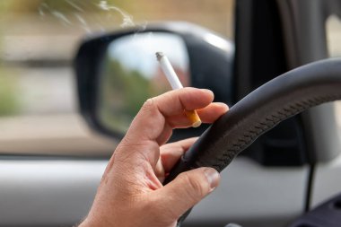 Close-up detail view of male hand driver holding cigarette while driving inside car on higway city street road on day time. Man person smoking inside vehicle. Low concentration attention danger.
