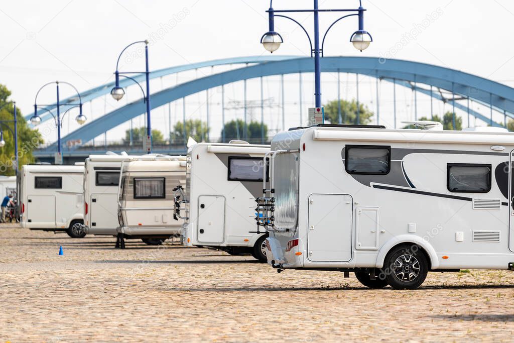 Many white modern campervan recreational motor home vehicles parked in row at camper park site Magdeburg city against Elbe river bridge. Motorhome campground stataion travel destination. RV lifestyle.