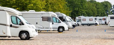 Many white modern campervan recreational motor home vehicles parked in row at camper park site Magdeburg city against Elbe river bridge. Motorhome campground stataion travel destination. RV lifestyle.