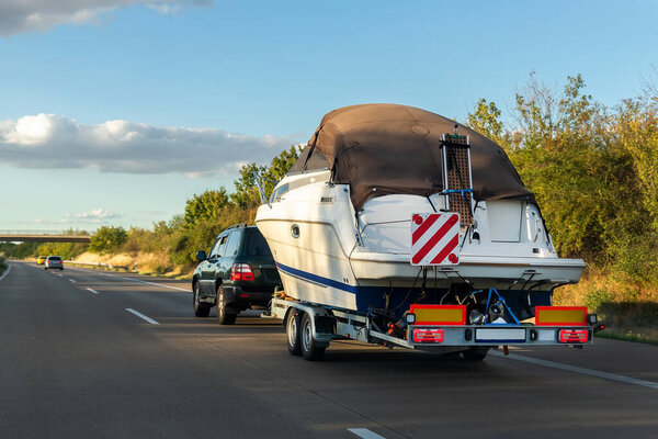 Big luxury cabin motorboat cruiser yacht trailer SUV carrier go to river lake on highway road sunset summer light. Luxury rich fishing leisure recreation lifestyle. Service maintenance transportation.