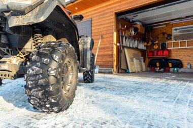 Back POV close-up detail view of quad bike offroad vehicle parked front house garage open door on sunny snowy cold winter day. ATV adventure extreme sport. Scoop snow removal tool eqiopment at home.