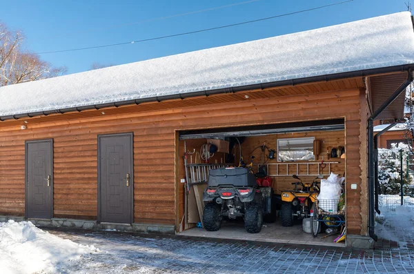 Facade view open door ATV home garage with quad bikes offroad vehicle parked sunny snowy cold winter day. ATV adventure extreme sport. House organized clutter warehouse tools equipment shed storage.