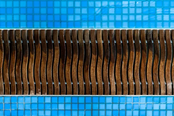 Close-up detail view of wet wooden pool edge overflow drain grain in blue mosaic tile. Abstract pattern of luxury poolside wood drainage grate. SPA water at hotel service and maintenance concept.