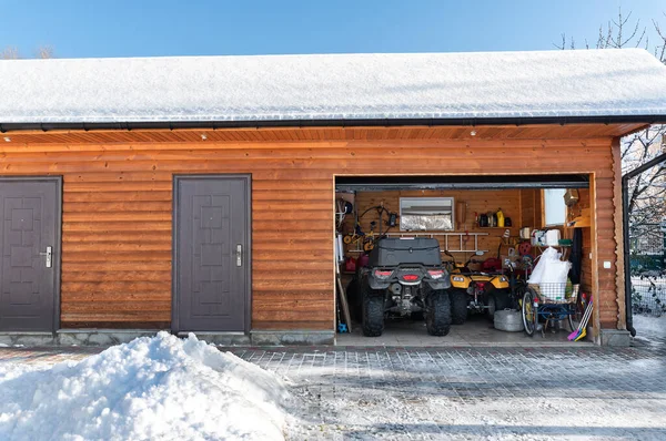 Facade view open door ATV home garage with quad bikes offroad vehicle parked sunny snowy cold winter day. ATV adventure extreme sport. House organized clutter warehouse tools equipment shed storage.