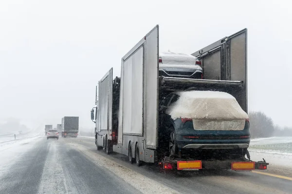 Tow truck car carrier semi trailer on highway carrying batch of new wrapped electric SUVs on motorway road at heavy snowfall in winter. Business distribution logistics service. Lorry driving highway.