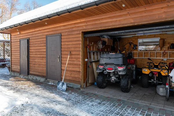 Facade view open door ATV home garage with quad bikes offroad vehicle parked sunny snowy cold winter day. ATV adventure extreme sport. House organized clutter warehouse tools equipment shed storage.