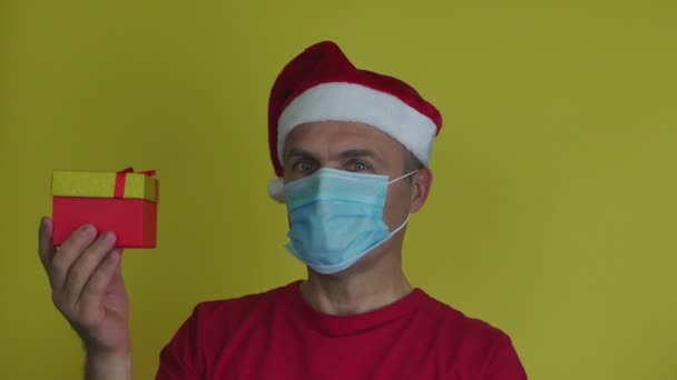 Un hombre con un sombrero y una máscara de Santa lleva una caja de regalo en la mano.. — Vídeo de stock