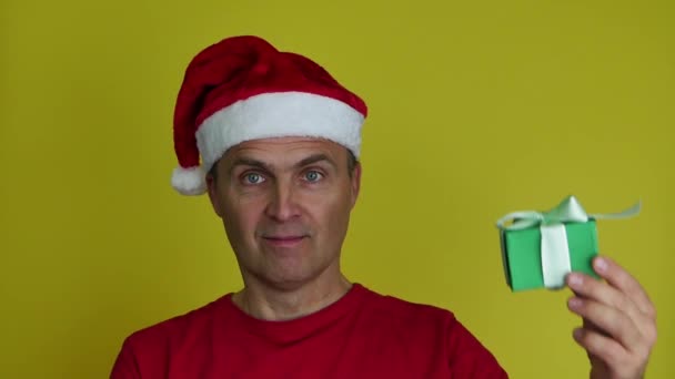 Een man in een kerstman hoed houdt een groene geschenkdoos met een strik in zijn hand. — Stockvideo